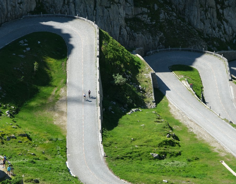Radfahrer erklimmen eine kurvenreiche Alpenstraße oberhalb eines Tals.